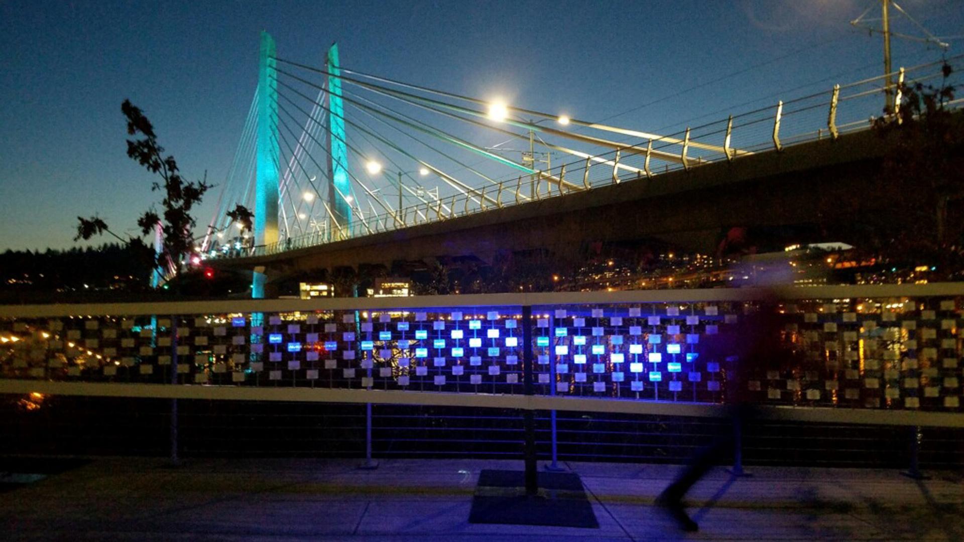 Night view of the Kinetic DeLight installation glowing blue on the Portlands waterfront with Tilikum Crossing in the background.